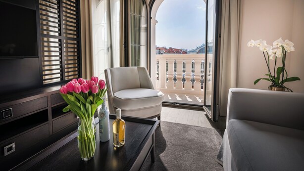 Habitación de hotel con televisión, un jarrón de tulipanes rosados, una botella, una silla blanca y vistas de edificios a través de grandes ventanales.