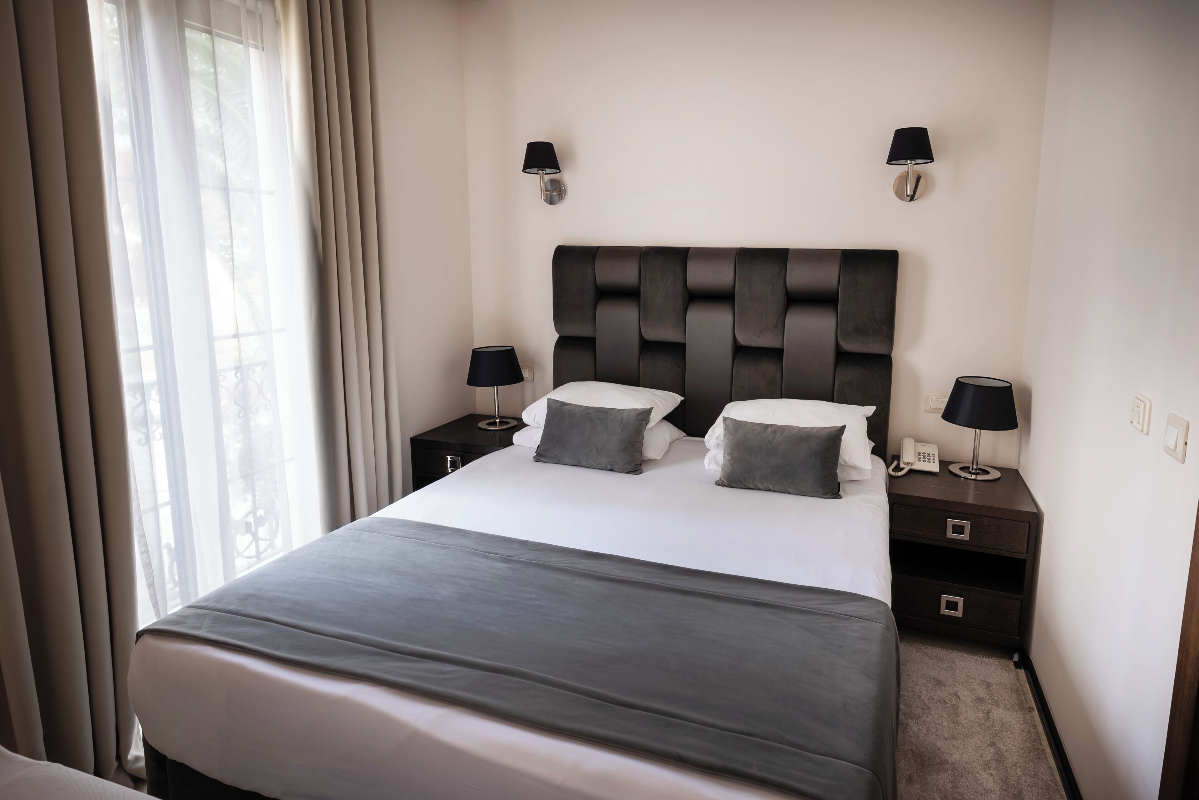 A modern hotel room with a large bed, two bedside tables, a headboard, and a window with curtains.