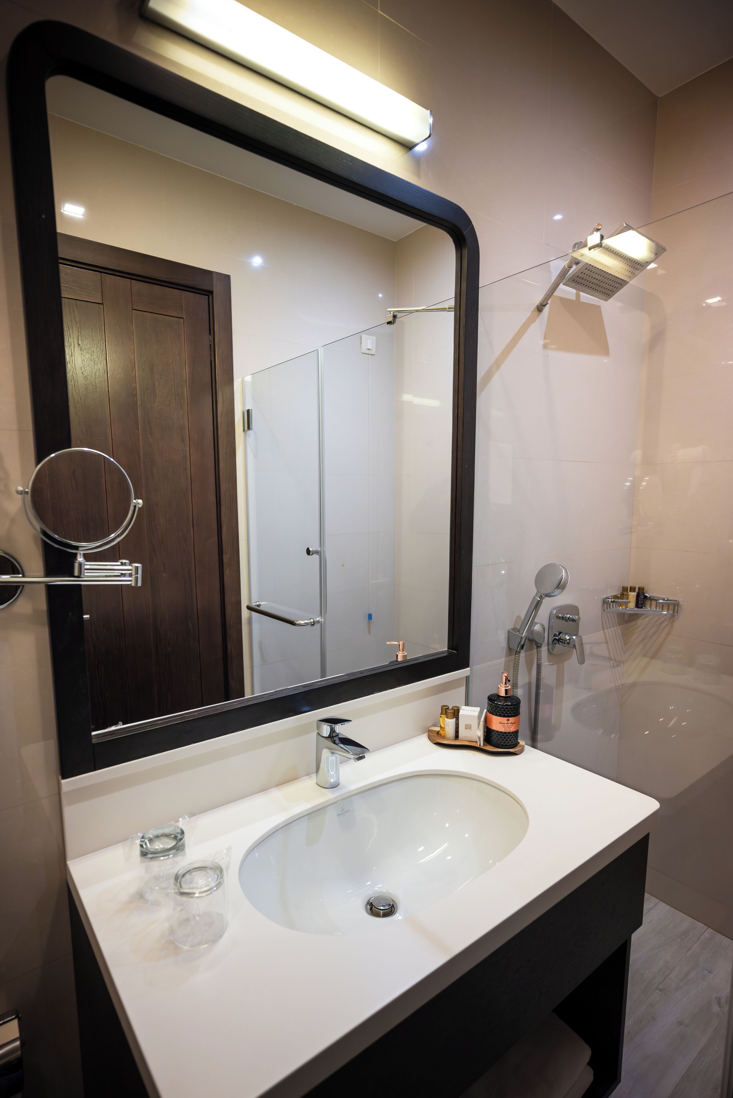 Un baño moderno con un espejo grande, un lavabo blanco, un cabezal de ducha manual y un perchero para toallas.