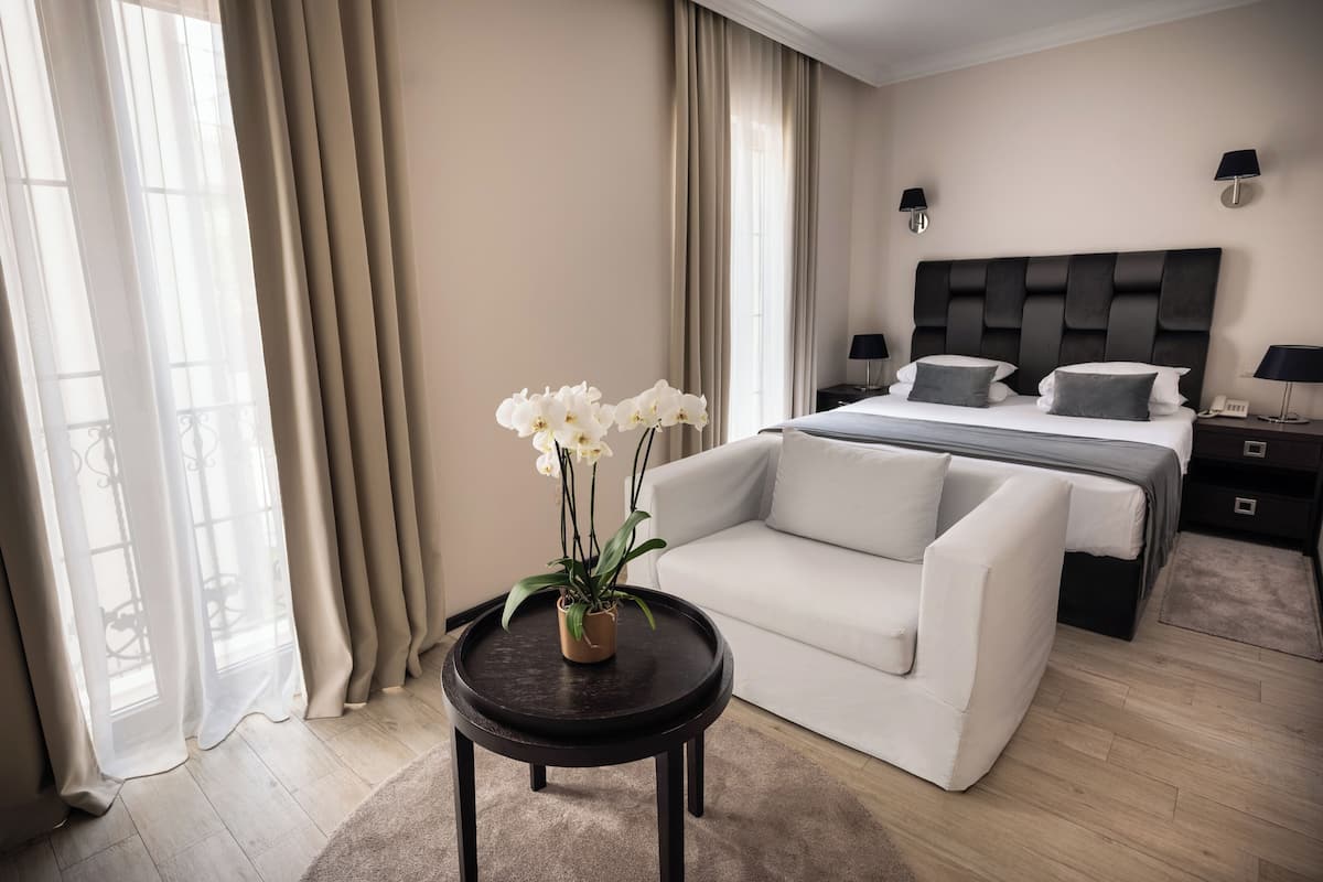 A modern hotel room with a bed, a sofa, a small table with flowers, and a view of the outdoors through large windows.