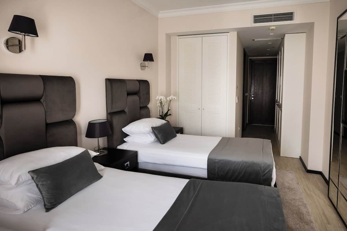 A hotel room with two beds, a nightstand, a lamp, a wardrobe, and a door leading to a hallway.