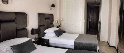 A hotel room with two beds, a nightstand, a lamp, a wardrobe, and a door leading to a hallway.