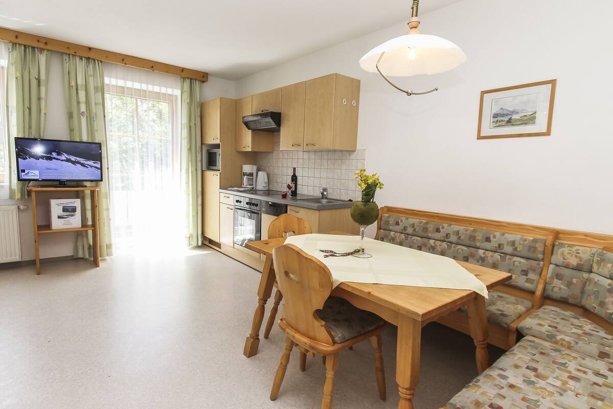 A modern kitchen with wooden cabinets, a dining table with chairs, a sofa, and a television on a stand.