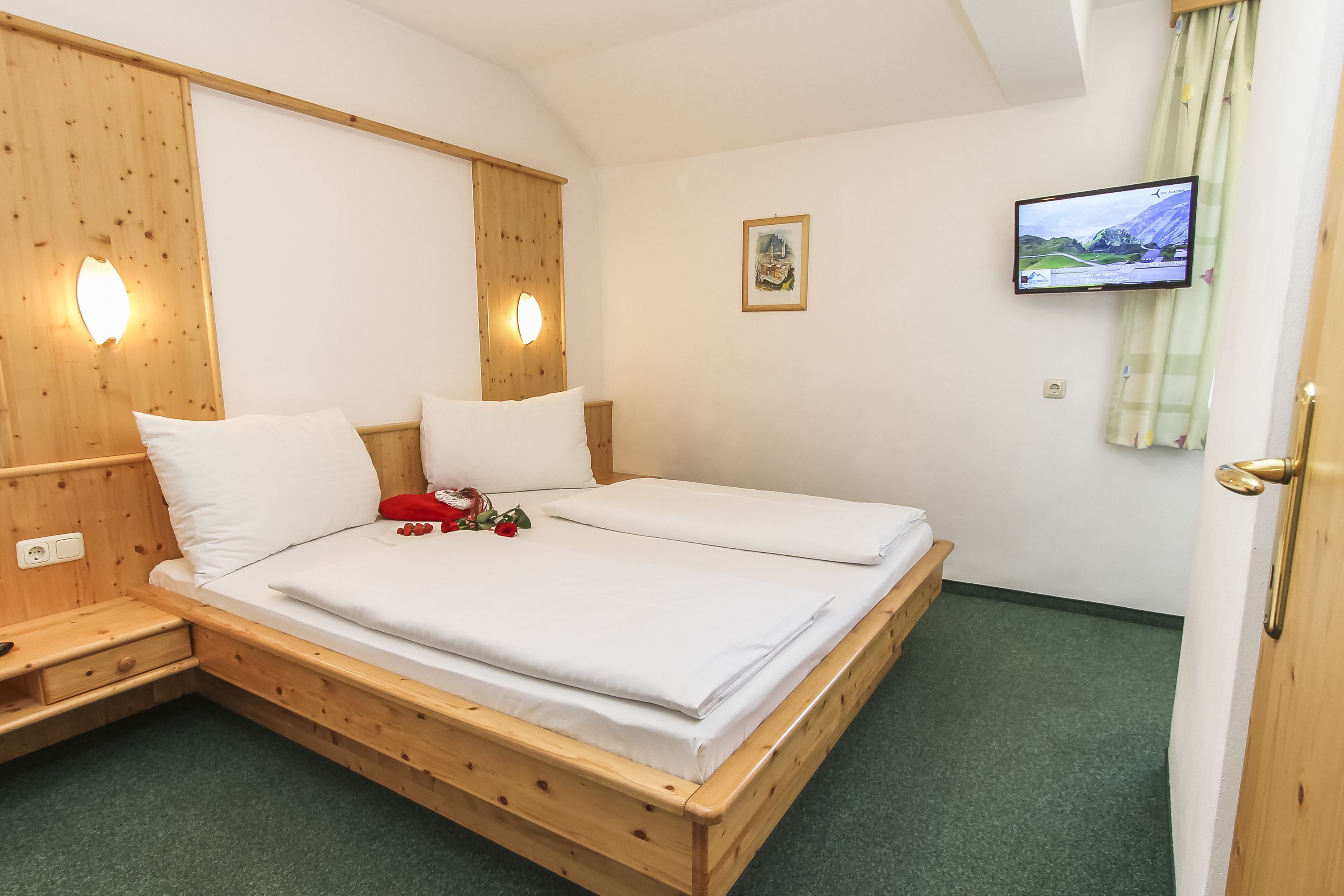 Una cama de madera con ropa de cama blanca, una mesita de noche, un televisor montado en la pared y un cuadro enmarcado.