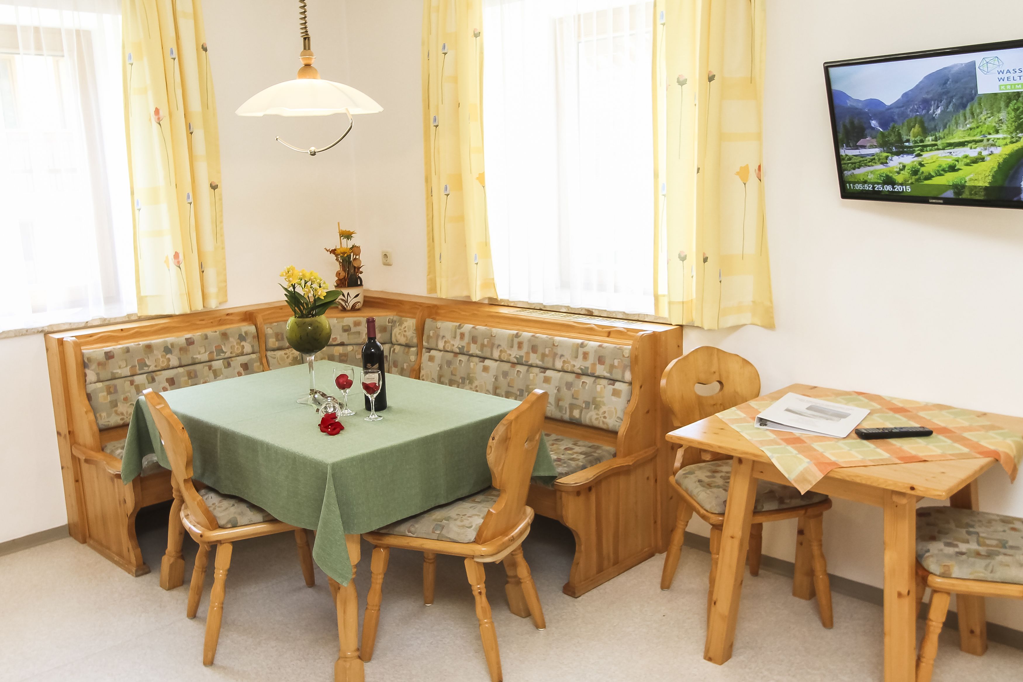 Un comedor con una mesa verde, sillas de madera, un banco con cojines estampados, un florero con flores y un televisor en la pared.