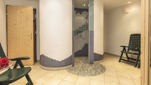 A spa room with a wooden door, a green chair, a small table with a red rose, and a shower area with blue-tiled walls. - Appartements Trinker (Zell am See)
