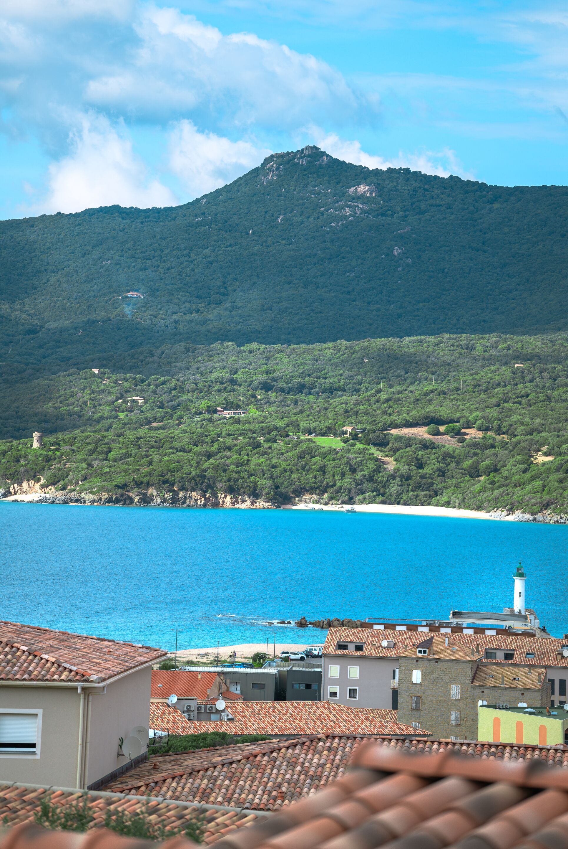 A coastal town with terracotta-roofed buildings, a lighthouse, and a mountain in the background.