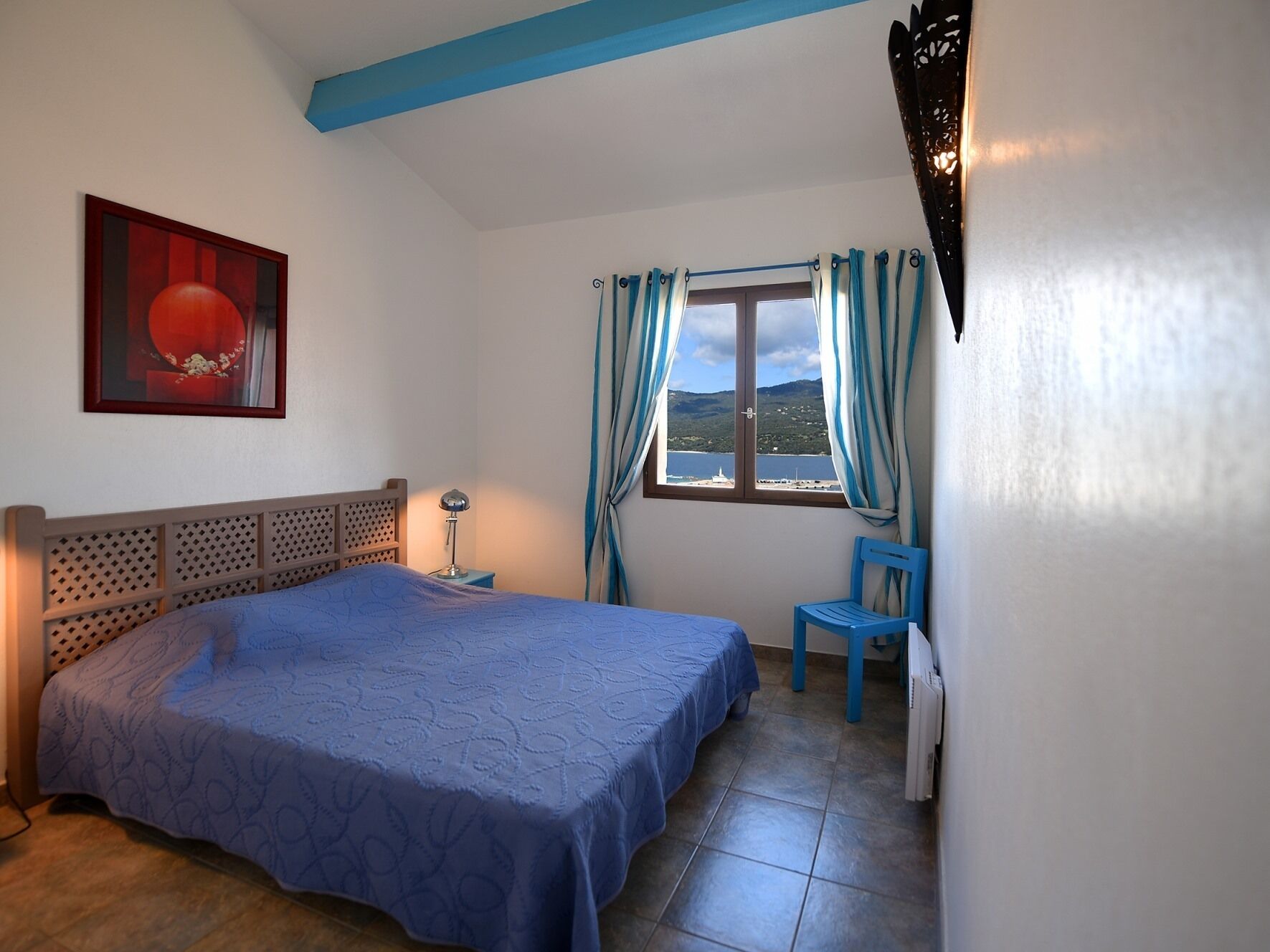 Une chambre à coucher comprenant un lit, une chaise, une table de chevet, une lampe, une fenêtre avec des rideaux et un tableau accroché au mur.