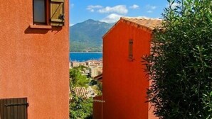 Un estrecho callejón con un edificio rojo, una planta en maceta y una vista de un pueblo costero y montañas al fondo.