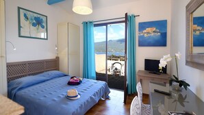 Una habitación de hotel con cama, escritorio con televisor, silla, mesa, balcón con vistas y obras de arte en las paredes.