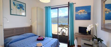 A hotel room with a bed, a desk with a TV, a chair, a table, a balcony with a view, and artwork on the walls.