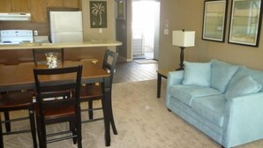 A living room with a beige carpet, a blue sofa, a dining table with chairs, and a kitchen area with wooden cabinets and a refrigerator. - Lake Arrowhead Drive (Myrtle Beach)