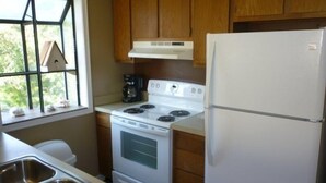 A compact kitchen with wooden cabinets, a white stove, a white refrigerator, and a window with a view of greenery. - Lake Arrowhead Drive (Myrtle Beach)