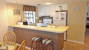 A compact kitchen with white cabinets, a stove, and a refrigerator. There are two bar stools and a dining table with chairs. - Lake Arrowhead Element (Myrtle Beach)