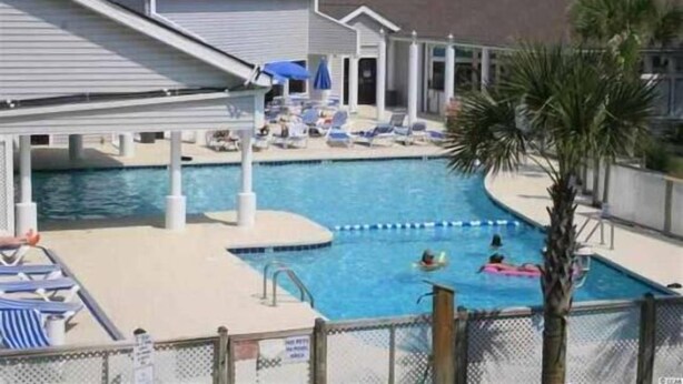 A swimming pool area with lounge chairs and umbrellas, surrounded by buildings. - Lake Arrowhead Turf (Myrtle Beach)