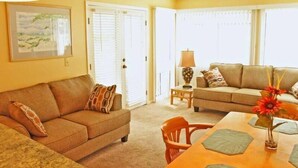 A living room with a beige sofa, a wooden dining table with placemats, and a vase with flowers. - Lake Arrowhead Turf (Myrtle Beach)