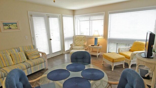 A living room with a striped sofa, round glass table, blue chairs, wicker chairs, and a TV on a side table. - Lake Arrowhead Break (Myrtle Beach)