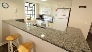 A kitchen with granite countertops, white cabinets, a microwave, stove, and a refrigerator. There are two wooden stools and a clock on the wall. - Lake Arrowhead Break (Myrtle Beach)