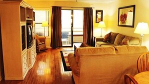 Una sala de estar con sofá, una silla, una mesa y un aparador. Hay una ventana con cortinas y una puerta que da a un espacio al aire libre.