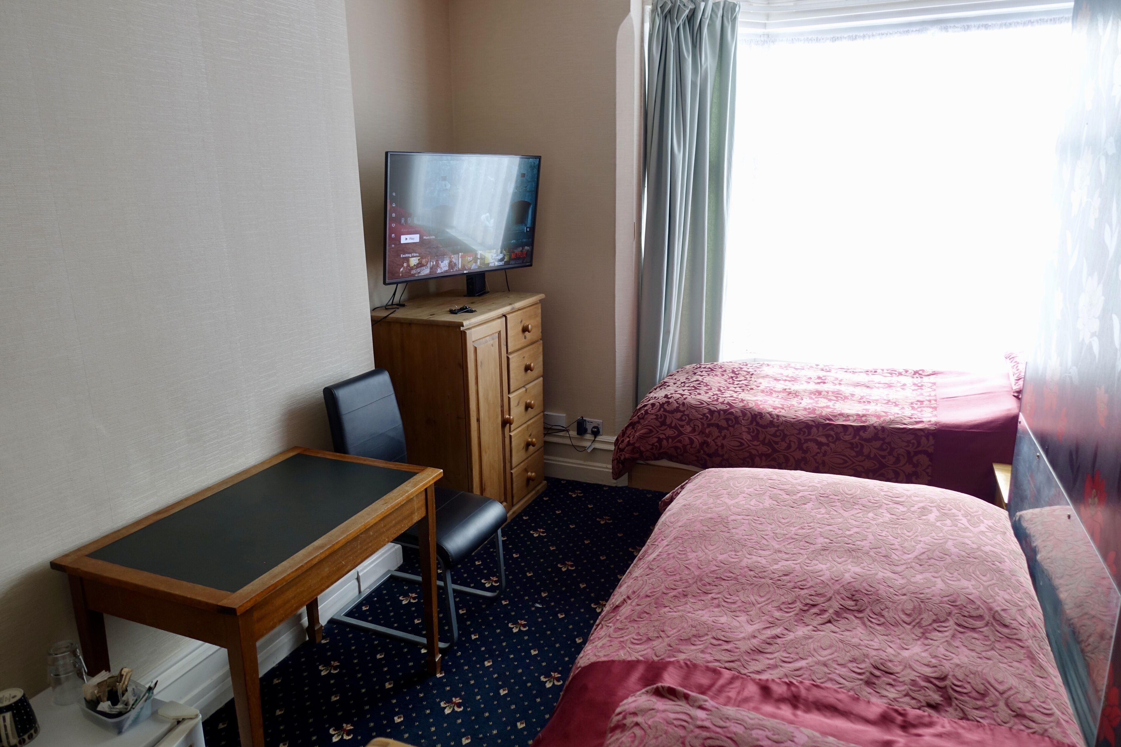 A hotel room with a bed, a desk with a chair, a television on a wooden dresser, and a window with curtains.