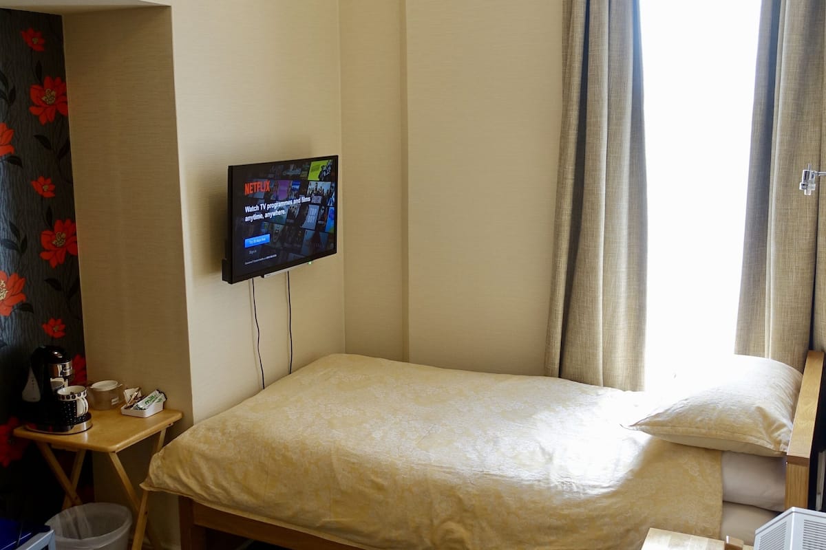 A hotel room with a bed, a desk with a computer, a TV mounted on the wall, and a ceiling light fixture.
