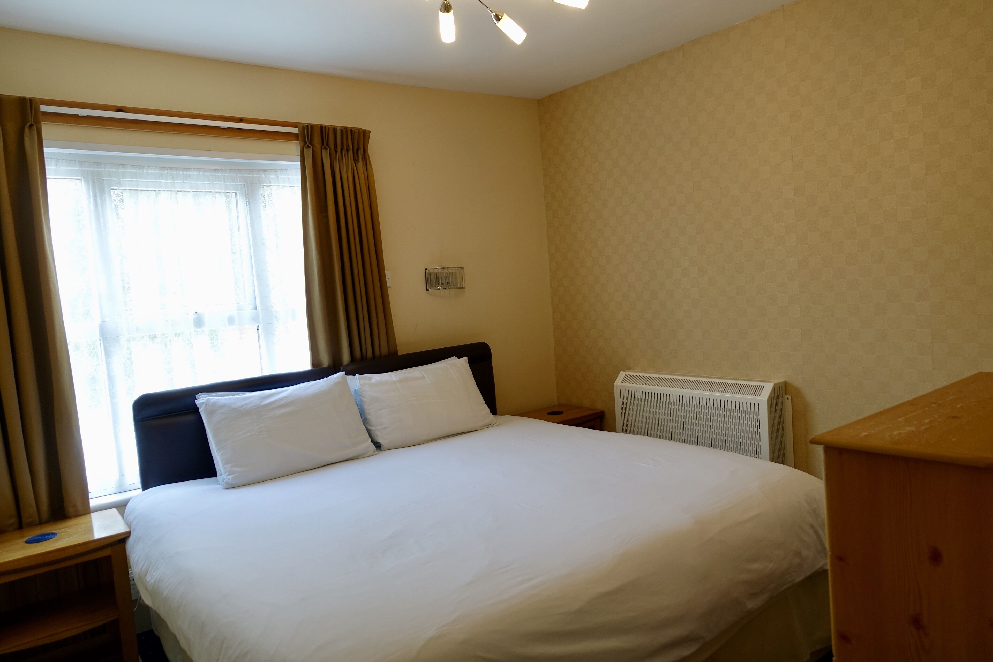 A hotel room with a large bed, a bedside table, a radiator, and a window with curtains.