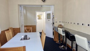 Quarto de hotel com uma mesa de jantar, cadeiras, área de cozinha com um banquinho e um relógio na parede.