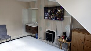 Quarto com cama, televisão na parede, armário de madeira, mesa pequena com cafeteira e banheiro com pia e espelho.