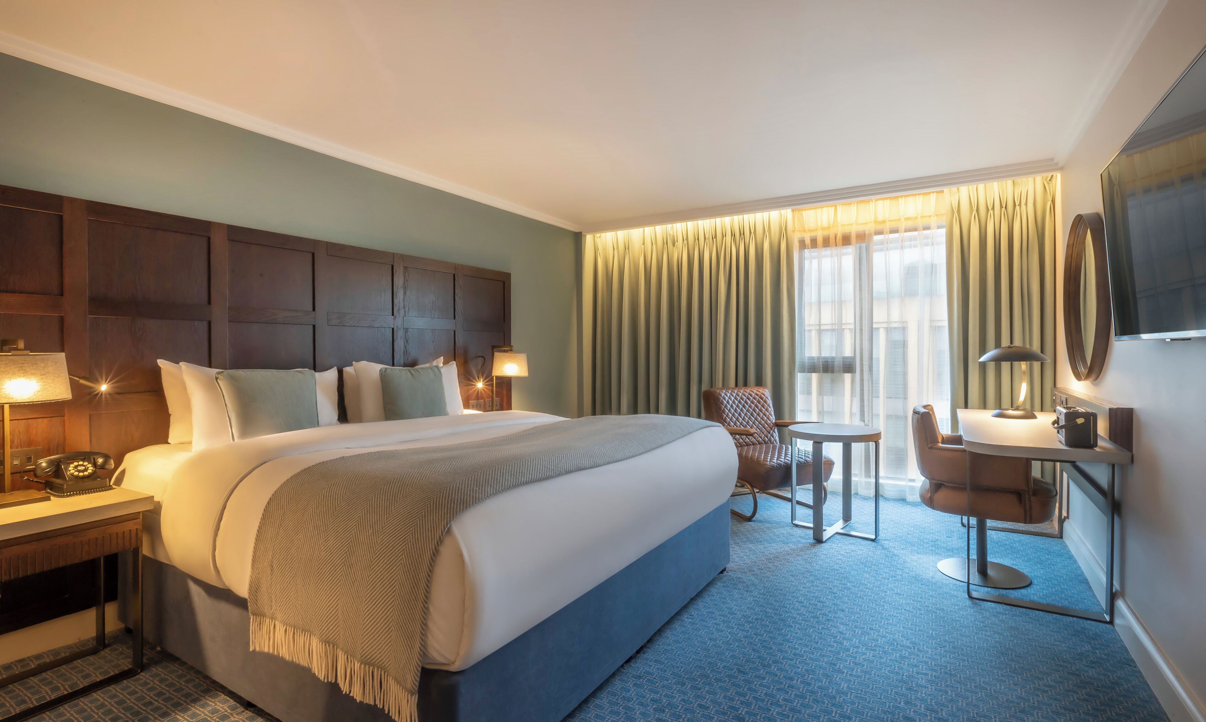 Une chambre d’hôtel avec un grand lit, un bureau avec un téléphone, une chaise, une petite table et un téléviseur fixé au mur.