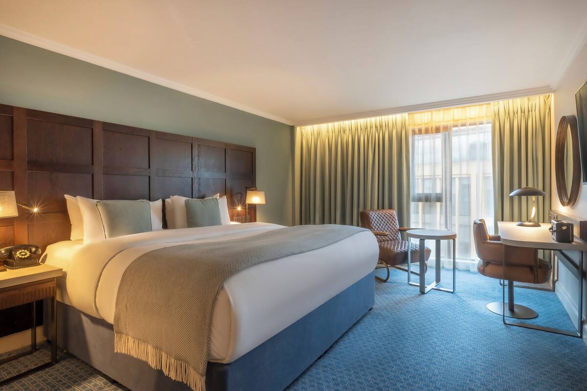 A hotel room with a large bed, a desk with a telephone, a chair, a small table, and a TV mounted on the wall.