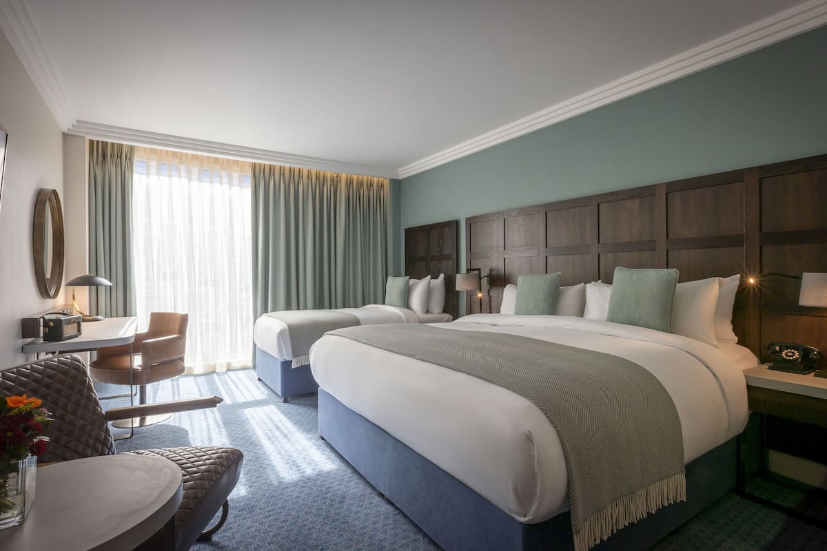 A hotel room with two beds, a desk, a chair, and a nightstand with a lamp.