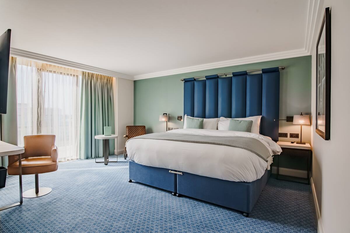 A hotel room with a large bed, a chair, a small table, and a nightstand with a lamp.