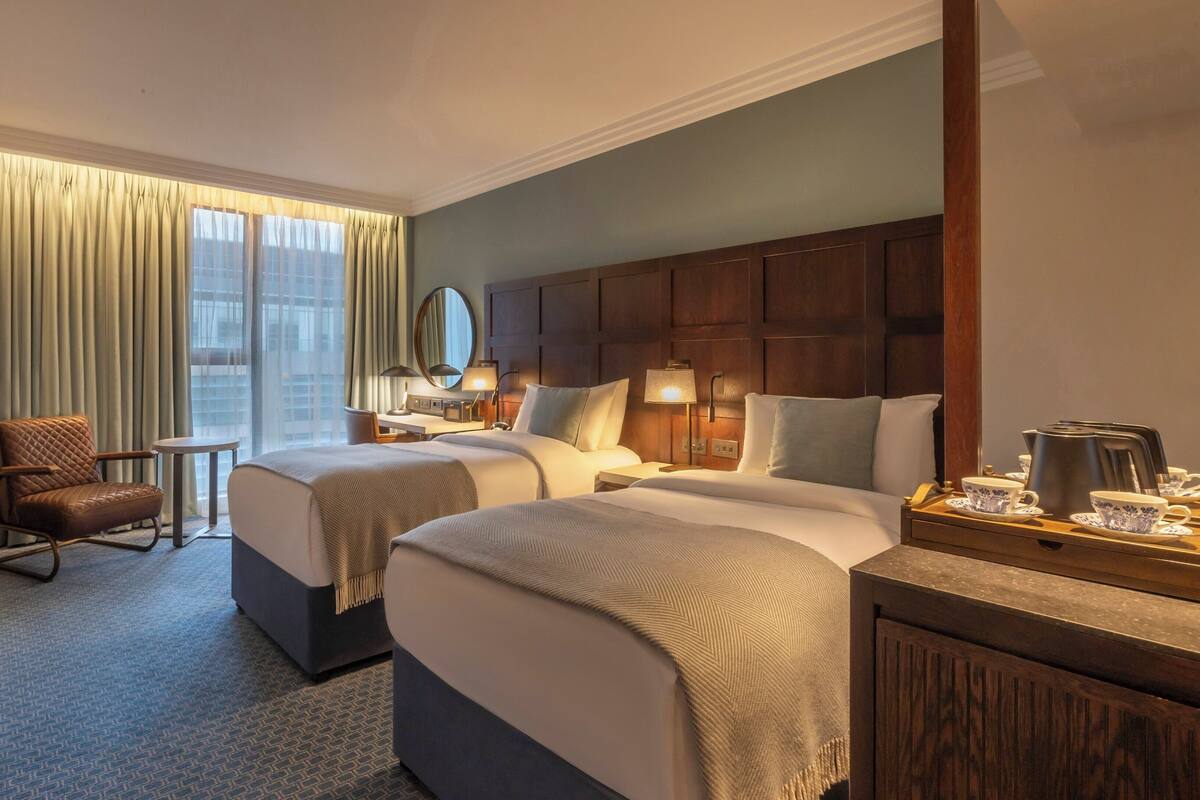 A hotel room with two beds, a desk, a chair, a TV, a window with curtains, and a coffee station.