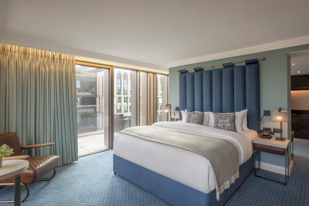 A modern hotel room with a large bed, a desk, and a chair. There is a sliding glass door leading to an outdoor area.