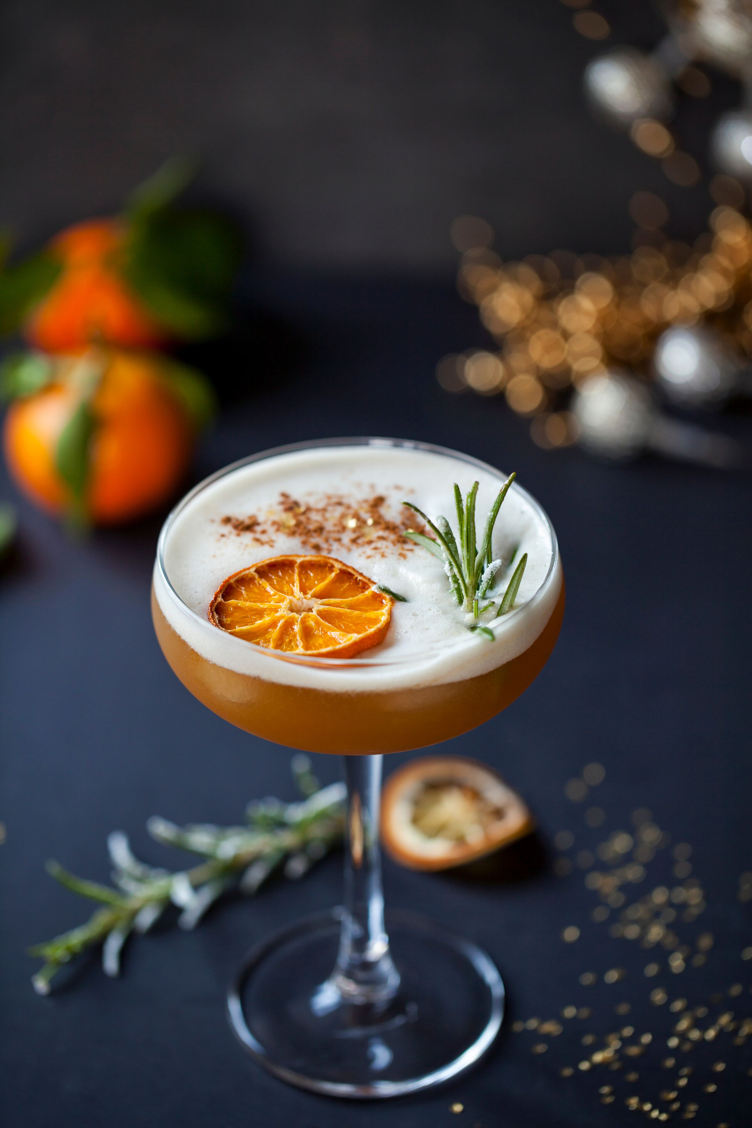 A cocktail in a stemmed glass with a slice of orange and a sprig of rosemary on top.
