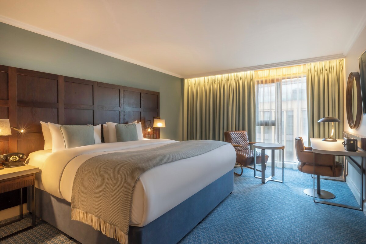 A hotel room with a large bed, a desk with a telephone, a chair, a small table, and a TV mounted on the wall.