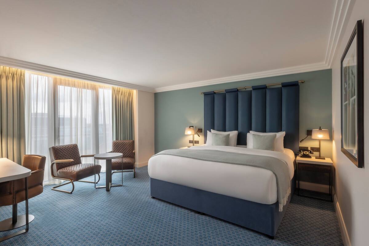A hotel room with a large bed, two chairs, a small table, and a nightstand with a lamp.