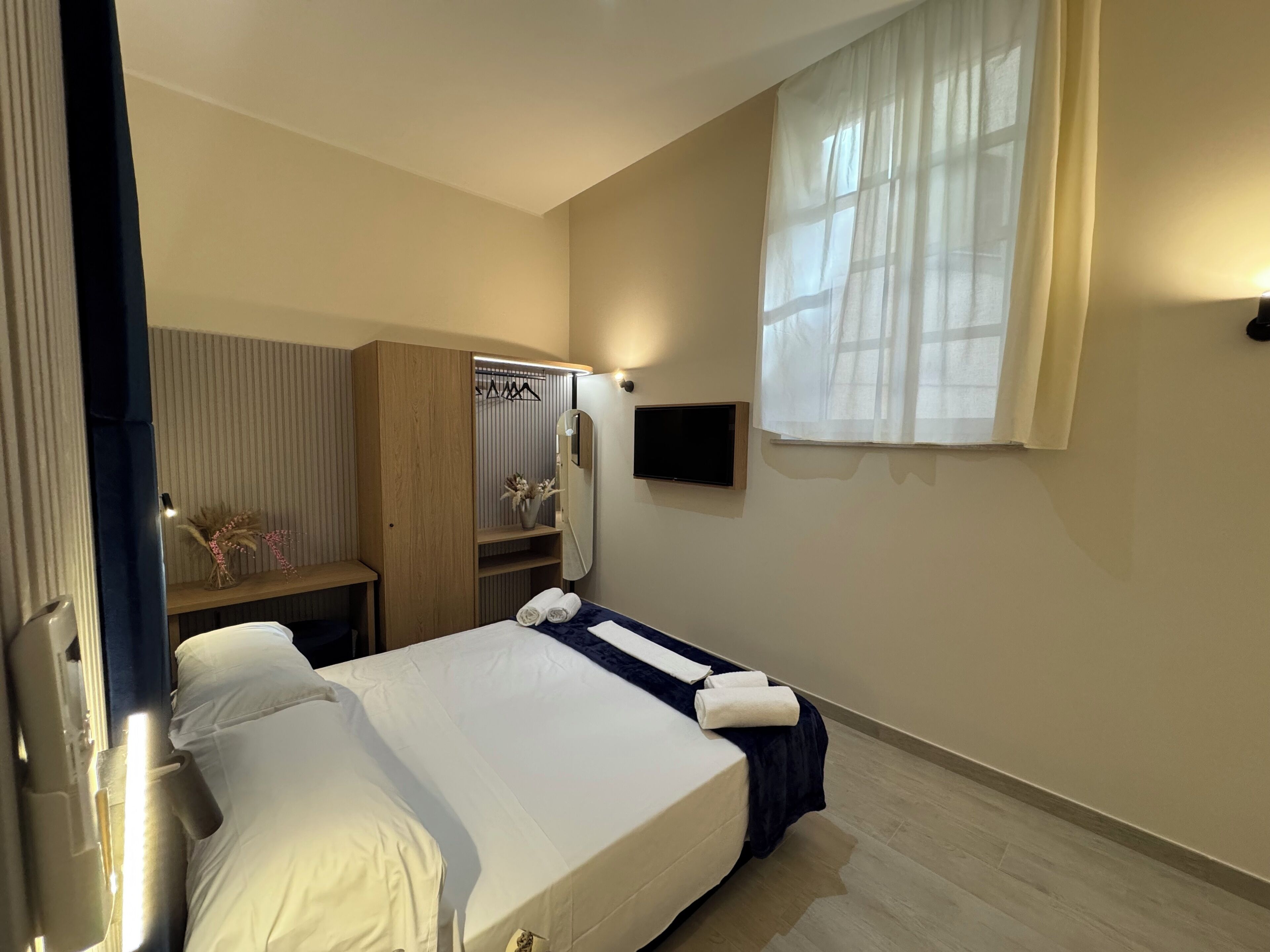 A hotel room with a bed, a wardrobe, a small table with a vase, and a window with curtains.