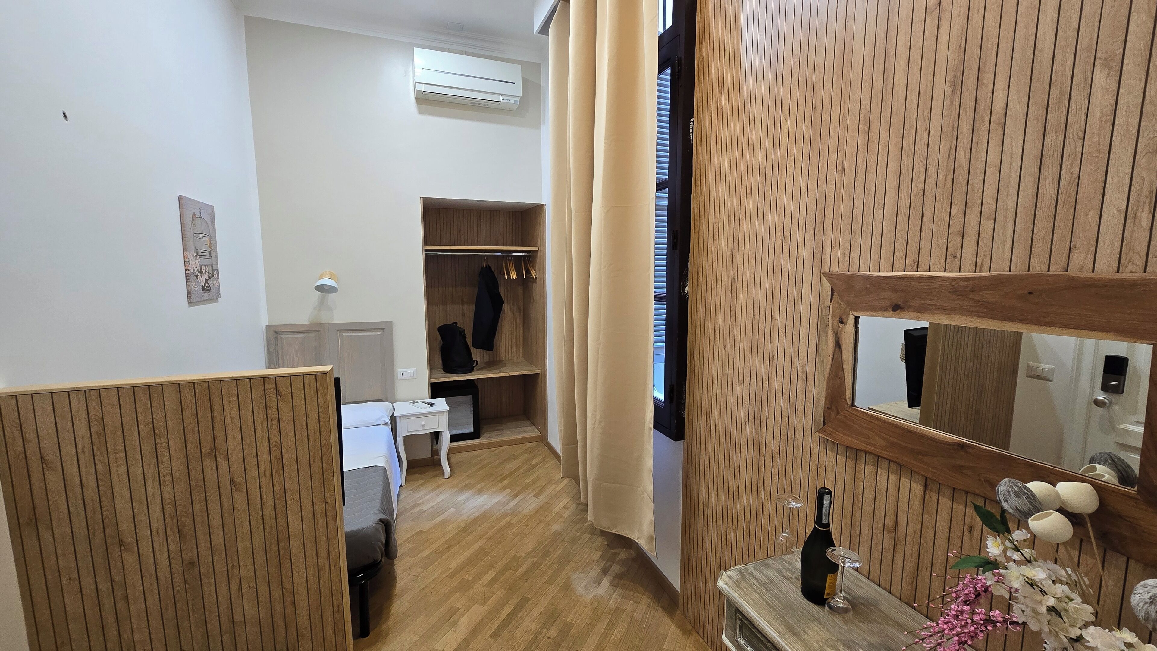 Una camera d'albergo con pavimento in legno, un letto, un comodino con una bottiglia e un bicchiere, un armadio e uno specchio.