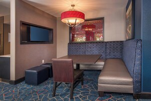 A hotel room with a dining area, a television mounted on the wall, a red pendant light, and a patterned carpet.
