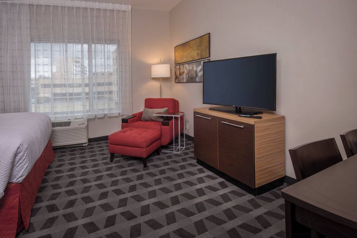 Ein Hotelzimmer mit einem Bett, einem roten Sessel, einem Fernseher, einem Holzschrank und einem gemusterten Teppich.