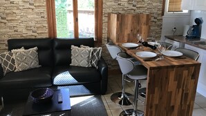 A modern living room with a black leather sofa, a wooden dining table set for four, and a kitchen area with white cabinets and a stone wall. - Les Plantous de Severo (Cenac-et-Saint-Julien)