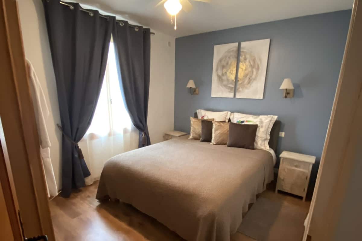Une chambre à coucher comprenant un lit, un ventilateur de plafond, une table de chevet, une lampe fixée au mur et une fenêtre avec des rideaux.