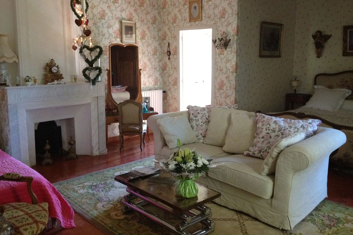 Ein gemütliches Wohnzimmer mit Kamin, floralem Wandpapier, einer Couch und einem Couchtisch mit Blumenvase.