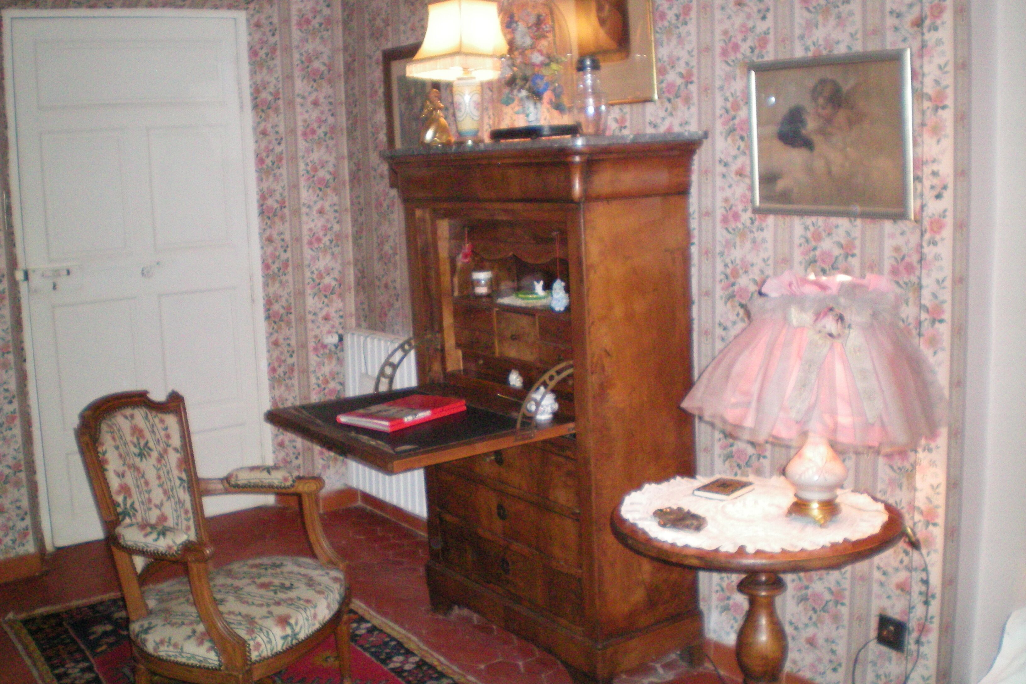 A room with a wooden cabinet, a chair, a table with a lamp, and floral wallpaper.