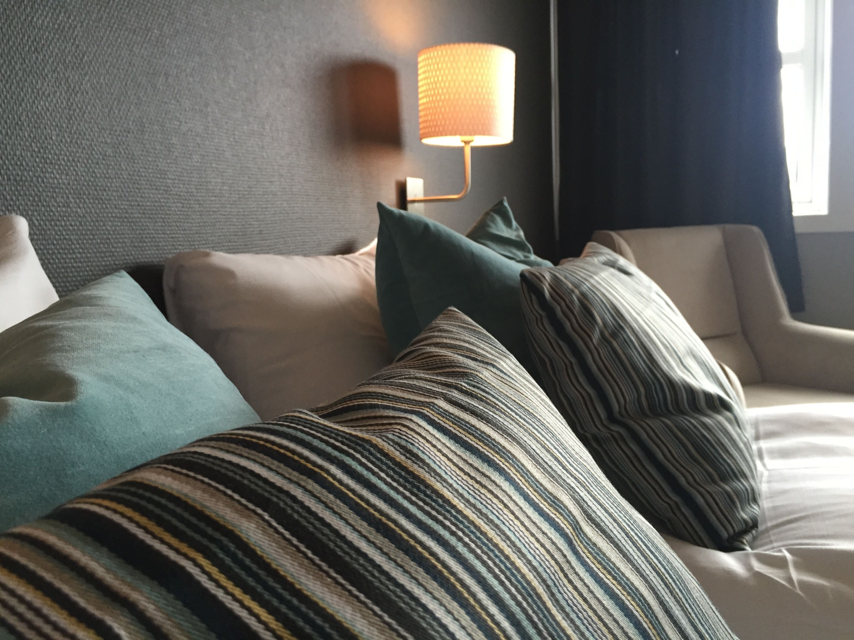 Et hotelværelse med en seng, puder, en lampe på væggen og et vindue med gardiner.