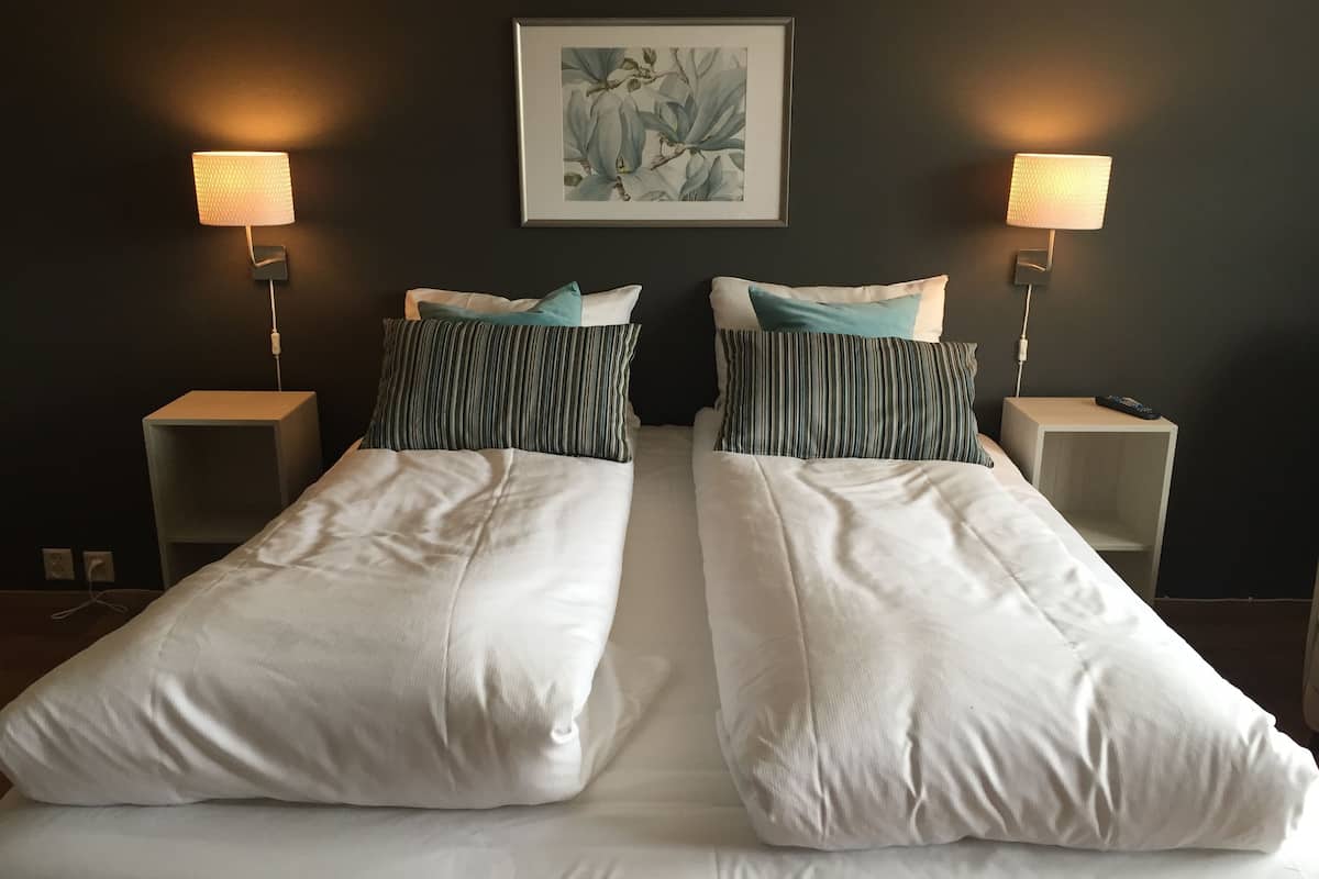 Un lit double avec du linge de lit blanc, deux tables de chevet avec des lampes et, au-dessus du lit, une œuvre d’art encadrée représentant des motifs floraux.