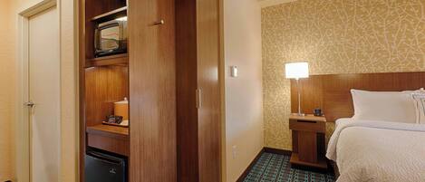 A hotel room with a bed, a wardrobe, a small refrigerator, a nightstand with a lamp, and patterned carpet.