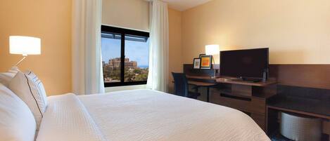 Habitación de hotel con una cama grande, un escritorio con televisor y vistas a un paisaje urbano a través de la ventana.