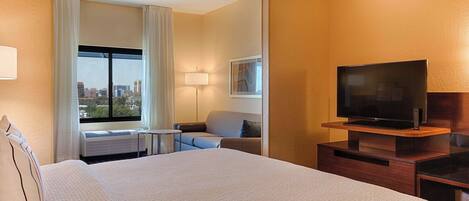 A hotel room with a large bed, a television on a wooden stand, a desk with a lamp, and a view of the city through a window.